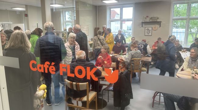 Foto gennem en rude med logoet Cafe FLOK på. Mange mennesker hygger og slapper af i cafeen med kaffe og mad.